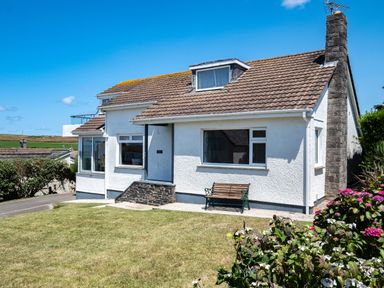 A white house with a tiled roof a bench on the front patio and a garden with flowers at Little Belz in Polzeath