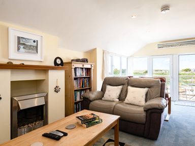 A living room with a leather sofa two cushions wooden coffee table books remote controls fireplace bookshelf and large windows at Little Belz in Polzeath