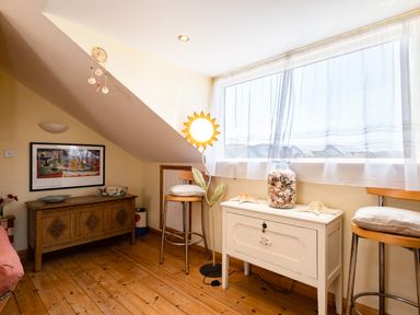 A room with wooden floor two stools a white chest with a jar of shells sun shaped lamp and a wooden cabinet with a framed picture at Little Belz in Polzeath