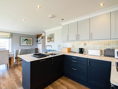 A kitchen with dark blue lower cabinets white upper cabinets and a dining area with chairs and a sideboard at Little Belz in Polzeath