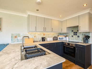 A kitchen with light grey upper cabinets dark lower cabinets a beige countertop sink oven and tiled backsplash at Little Belz in Polzeath