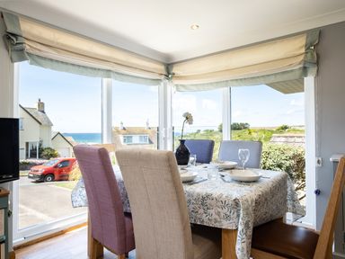 A dining room with four chairs around a table set with bowls and glasses by large windows showing houses and a sea view at Little Belz in Polzeath
