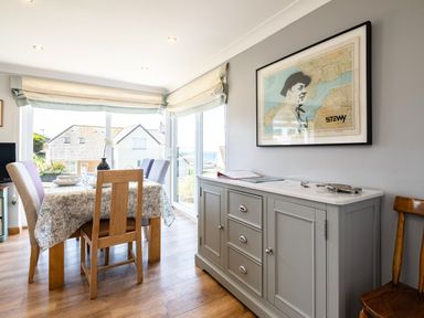 A dining area with a wooden table and chairs next to large windows and a sideboard with framed artwork on the wall at Little Belz in Polzeath
