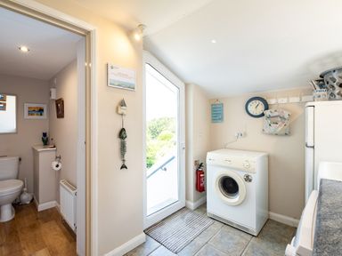 A laundry room with a washing machine a door leading outside a clock on the wall and a bathroom visible through an open door at Little Belz Polzeath
