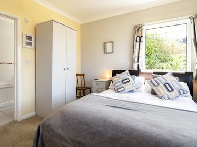 A bedroom with a double bed, bedside table with lamp, wardrobe, wooden chair and window with curtains at Little Belz in Polzeath