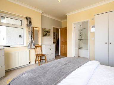 A bedroom with a bed a wooden chair a chest of drawers a window wardrobe and an en suite bathroom at Little Belz in Polzeath