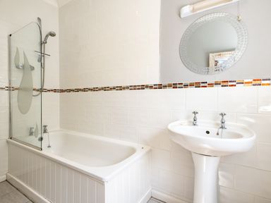 A bathroom with a bathtub shower combo a glass screen a tiled wall and a pedestal sink with a mirror above at Little Belz in Polzeath