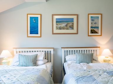 A bedroom with two single beds with striped pillows framed pictures above and bedside lamps at Little Belz in Polzeath