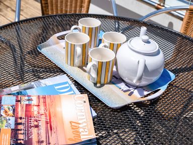 A round metal mesh outdoor table with a tray holding a white teapot and four striped mugs next to magazines at Little Belz in Polzeath