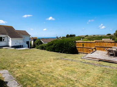 A backyard with grass lawn a white house a wooden deck with chairs and table and a view of the sea at Little Belz in Polzeath