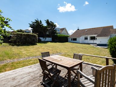 A wooden table with four chairs on a wooden deck overlooking a grassy backyard with bushes and a white house at Little Belz in Polzeath