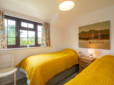 A bedroom with two single beds with yellow bedspreads a wooden nightstand with a lamp a window with floral curtains and a wall hanging of boats at Hazelhurst in Rock