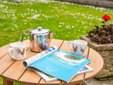 A wooden outdoor table with two rooster mugs a metal teapot and magazines on a lawn with flowers at Hazelhurst Rock