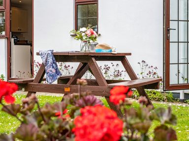 A wooden picnic table with a hat flowers and fabric on it outside a white house with open doors and red flowers in the garden at Hazelhurst in Rock