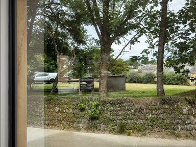 View through a window of trees a stone wall a grass lawn a parked white vehicle and a sign that reads Porthilly Farm Private Property in Rock