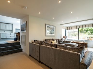 A living room with a sectional sofa, cushions, framed artwork on the wall, a lamp on a wooden console table under a large window at Porthilly Greys in Rock