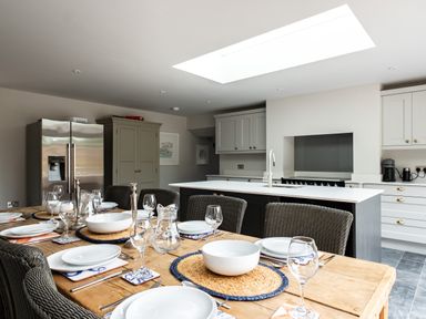 A kitchen with a dining table set with plates glasses and cutlery next to a kitchen island with a sink at Porthilly Greys in Rock