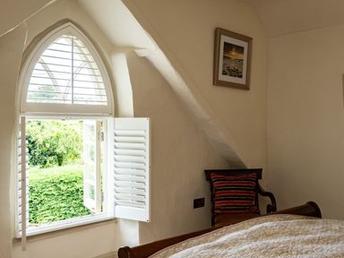 A bedroom with an arched window with shutters a chair with a striped cushion and a framed picture on the wall at Porthilly Greys in Rock