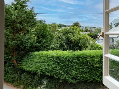 A view of green trees, shrubs, and rooftops seen through an open window at Porthilly Greys in Rock