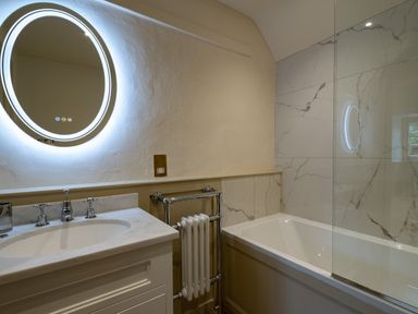 A bathroom with a round illuminated mirror above a sink and a bathtub with marble tiled walls at Porthilly Greys in Rock