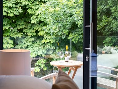 A small wooden table with a bottle of wine and two wine glasses on a balcony with wooden chairs and green trees outside Porthilly Greys in Rock