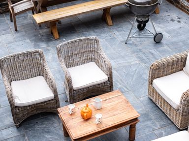An outdoor seating area with three wicker chairs around a wooden table with a teapot and cups and a charcoal grill on a tiled floor