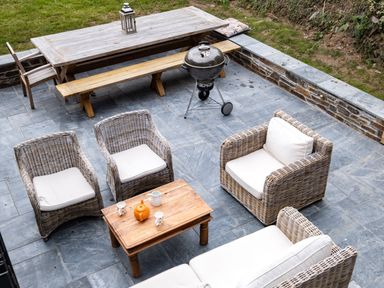 An outdoor patio with wicker chairs a wooden table a wooden bench a metal grill and grass at Porthilly Greys in Rock