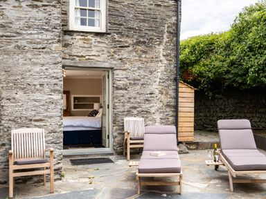 A stone patio with wooden chairs and cushioned loungers outside a stone house with an open door showing a bedroom at Porthilly Greys in Rock