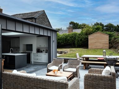 An outdoor patio area with wicker chairs and a wooden table next to an open kitchen and a garden shed in the background at Porthilly Greys in Rock