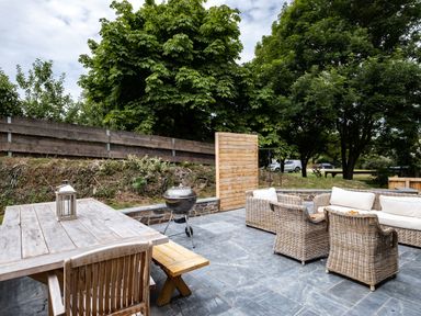 An outdoor patio with a wooden dining table and chairs a round charcoal grill and wicker seating with cushions surrounded by trees at Porthilly Greys in Rock
