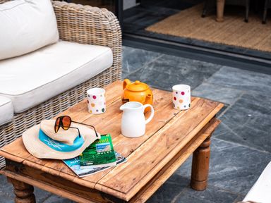 A wooden coffee table with a hat, sunglasses, magazines, teapot, jug, and two polka dot mugs next to a wicker sofa at Porthilly Greys in Rock