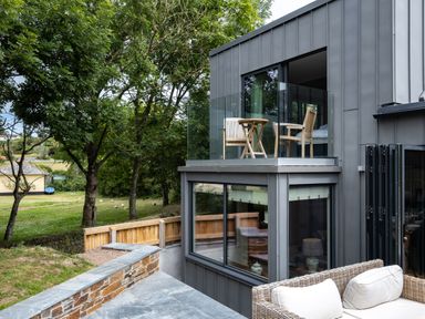 An outdoor patio with wicker seating and cushions next to a modern building with glass balcony and wooden chairs at Porthilly Greys in Rock