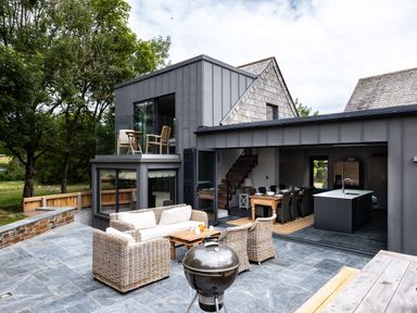 An outdoor patio with wicker furniture and a barbecue grill next to a modern house with an open kitchen and dining area at Porthilly Greys in Rock