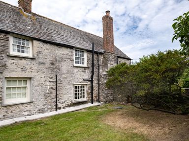 An exterior view of a stone house with white windows a brick chimney and a garden with grass and a tree at Porthilly Greys in Rock