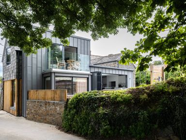 A modern two-story house with glass balcony and open kitchen doors surrounded by trees at Porthilly Greys in Rock