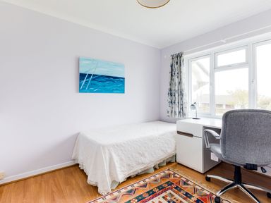A small bedroom with a bed, white desk with a lamp, gray office chair, window with curtains and a painting of the sea on the wall at Trebartha Daymer Bay
