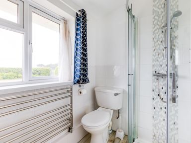 A bathroom with a toilet, glass shower, window with curtain, toilet paper holder and towel rack at Trebartha in Daymer Bay