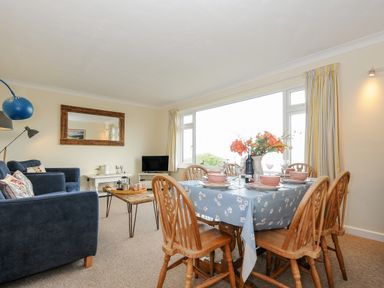 A dining table with six wooden chairs set with dishes and flowers next to a sitting area with blue sofas and a coffee table at Trevega Polzeath