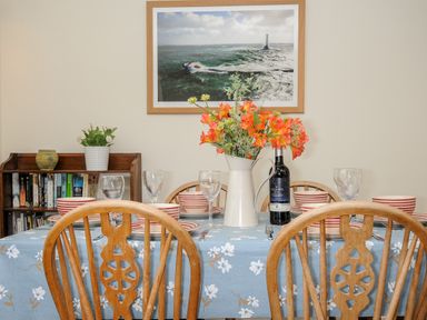 A dining table with a blue floral tablecloth set with striped bowls and glasses with a vase of orange flowers and a bottle of wine a bookshelf with books and plants and a framed ocean photo on the wall at Trevega in Polzeath