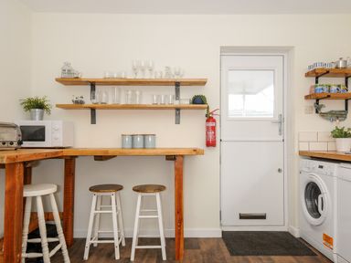 A kitchen corner with wooden shelves holding glasses and mugs a wooden counter with three stools a microwave a washing machine and a white door at Trevega Polzeath