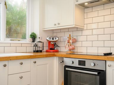 A kitchen corner with a wooden countertop a red coffee machine striped mugs on a wooden mug tree a black oven white cabinets and a window with plants visible outside at Trevega in Polzeath