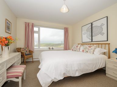 A bedroom with a double bed white bedding floral pillows a wooden chair and a dressing table with flowers at Trevega Polzeath