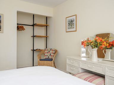 A bedroom with a wicker chair with a cushion an open closet with hangers and shelves a white dresser with a mirror and flowers at Trevega Polzeath