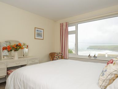 A bedroom with a white bed, a dressing table with flowers, a wicker chair, and a large window showing a sea view at Trevega in Polzeath