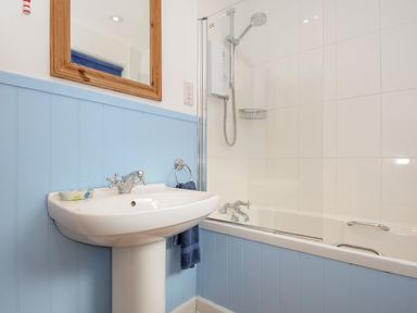 A bathroom with a white sink, wooden framed mirror, blue wall paneling, and a bathtub with a shower at Trevega Polzeath