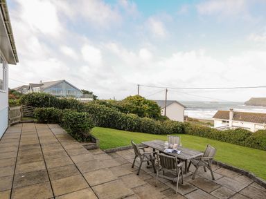 A patio with wooden table and chairs overlooking a grassy garden and houses with the sea in the background at Trevega in Polzeath