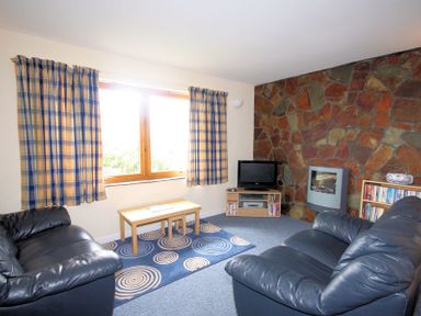 A living room with two black leather sofas a wooden coffee table a TV on a stand a stone wall and checked curtains at Polzeath Court 4 in Polzeath
