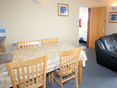 A dining area with a wooden table covered with a floral tablecloth and four wooden chairs next to a black leather sofa in a room at Polzeath Court 4 in Polzeath
