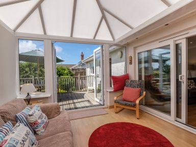 A conservatory with a sofa and cushions a wooden chair with cushions a round red rug and patio doors leading to an outdoor deck with a table umbrella at Cogenhoe in Rock