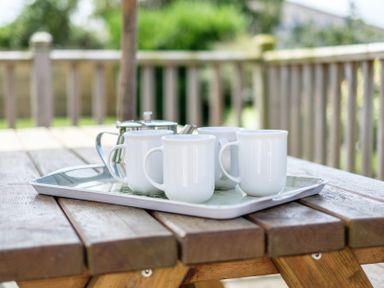 A wooden outdoor table with four white mugs and a metal teapot on a tray with a wooden fence in the background at Cogenhoe Rock